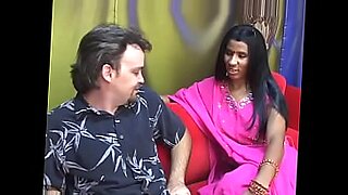 Young Indian woman gives blow job to older man on red couch.